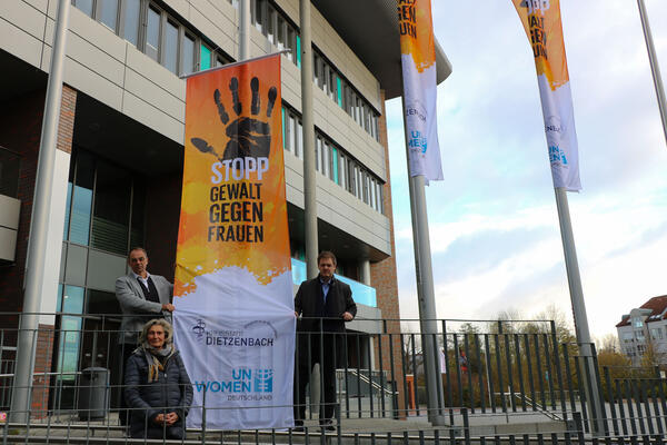 Dietzenbachs Frauen- und Gleichstellungsbeauftrage sowie Bürgermeister und Erster Stadtrat halten vor dem Rathaus die orangefarbene Flagge hoch mit der Aufschrift "Stopp Gewalt gegen Frauen"