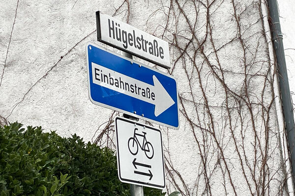 Straßenschild Hügelstraße, gekennzeichnet als Einbahnstraße, darunter ein Schild mit Fahrrad und Pfeilen in beide Richtungen