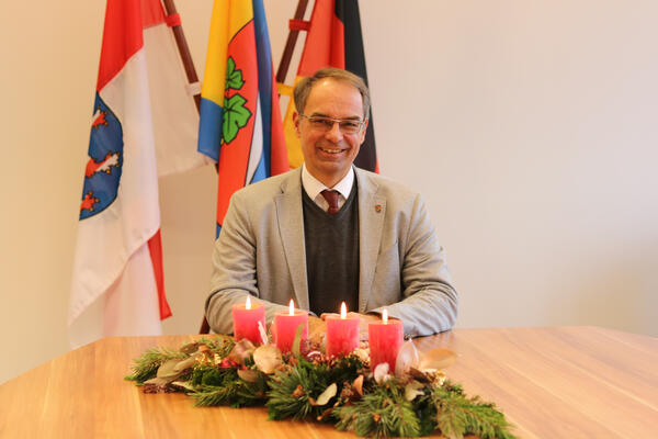 Ein Mann, der Bürgermeister, sitzt am Tisch, vor im ein Adventskranz mit vier Kerzen, hinter ihm die Flaggen von Dietzenbach, Hessen sowie Deutschland
