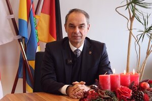 Bild vergr&ouml;&szlig;ern: Ein Mann sitzt am Tisch hinter einem festlichen Gesteck aus Zweigen und roten Kerzen.