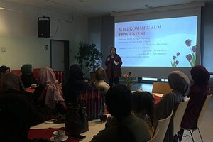Bild vergr&ouml;&szlig;ern: In einem Saal vor einer Leinwand-Pr&auml;sentation sitzen Frauen und M&auml;dchen an Tischen, eine Frau spricht vor der Gruppe.