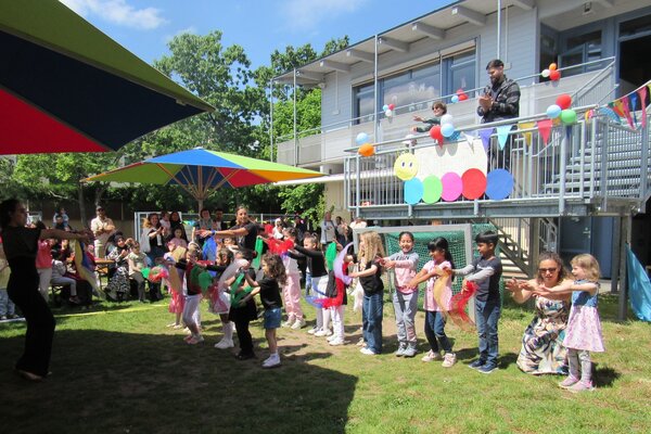 Viele Kinder stehen auf einer Wiese vor einem Geb&auml;ude und Tanzen mit Eltern und Erziehern.