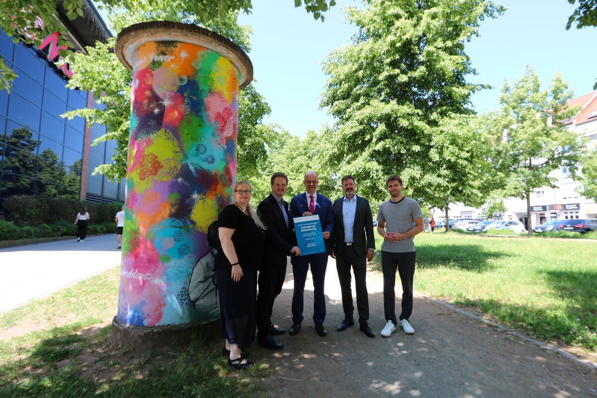 Eine Frau und vier M&auml;nner stehen vor einer bunt bemalten Litfa&szlig;s&auml;ule und halten symbolisch eine Urkunde zur F&ouml;rderung des Projekts "Demokratie leben" in den H&auml;nden.