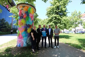 Bild vergr&ouml;&szlig;ern: Eine Frau und vier M&auml;nner stehen vor einer bunt bemalten Litfa&szlig;s&auml;ule und halten symbolisch eine Urkunde zur F&ouml;rderung des Projekts "Demokratie leben" in den H&auml;nden.
