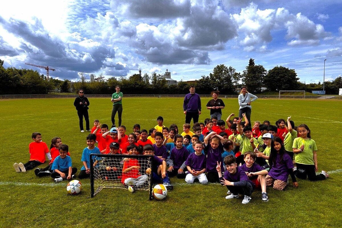 Viele Kinder, M&auml;dchen und Jungen sitzen in bunten Trikots auf einer Wiese vor einem kleinen Fu&szlig;balltor.