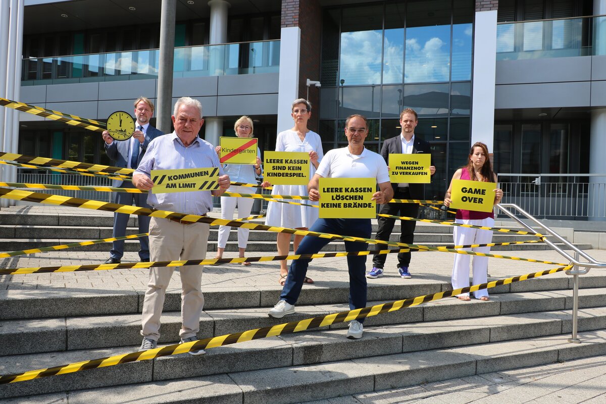 M&auml;nner und Frauen stehen zwischen gelb-schwarz-gestreiftem Flatterband vor einem Geb&auml;ude und halten Schilder und Transparente in den H&auml;nden.