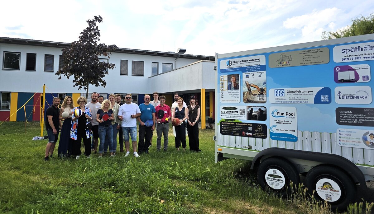 M&auml;nner und Frauen auf einer Wiese neben einem Transportanh&auml;nger. Auf diesem sind Firmennamen abgedruckt und die Personen halten teilweise Spielger&auml;te in den H&auml;nden.