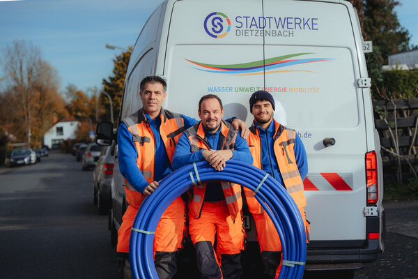 Drei M&auml;nner in Arbeitsanz&uuml;gen vor einem Transporter mit der Aufschrift "Stadtwerke Dietzenbach". Vor ihnen eine aufgerollte Wasserleitung.