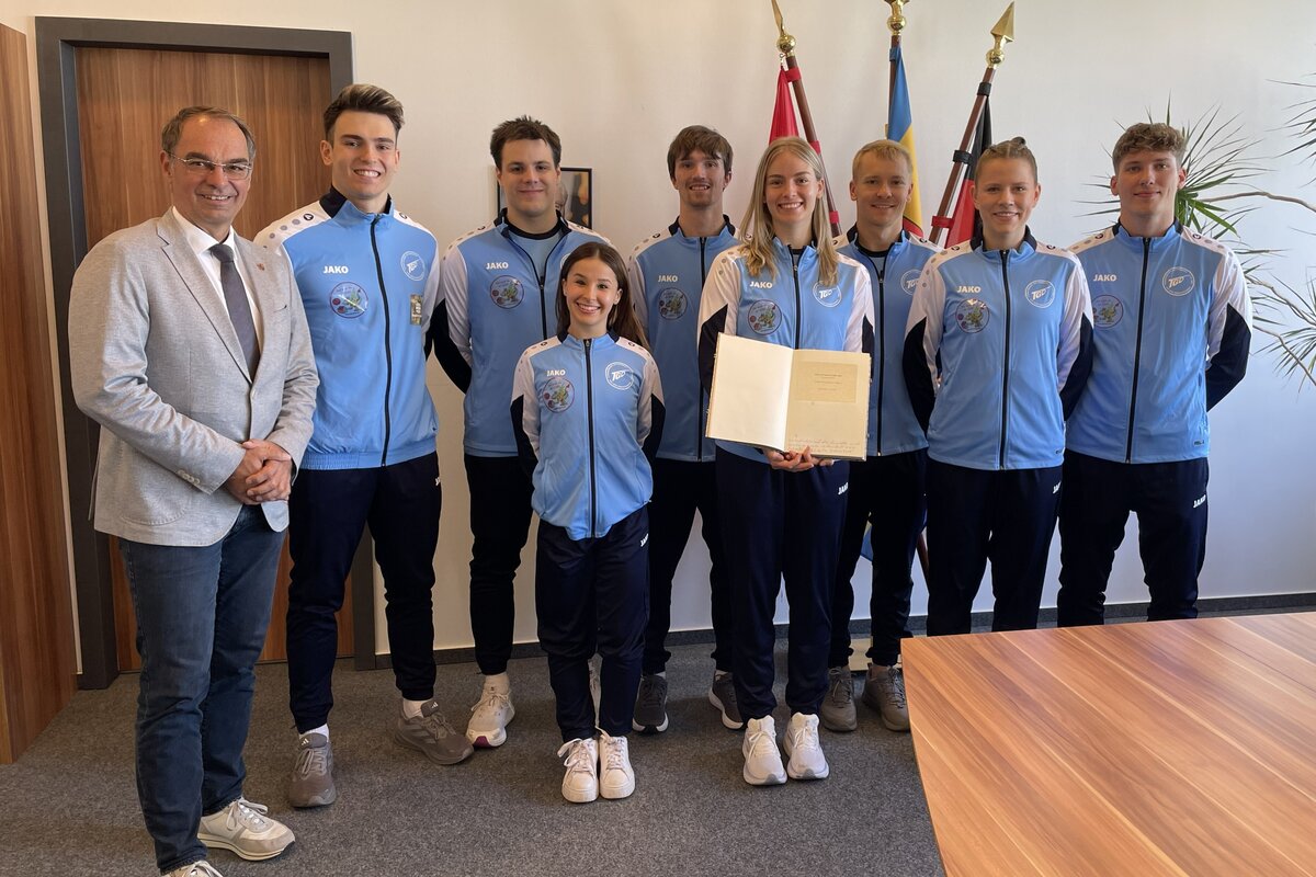 Junge Frauen und M&auml;nner in Trainingsanz&uuml;gen, eine von ihnen h&auml;lt ein aufgeschlagenes Buch in den H&auml;nden.
