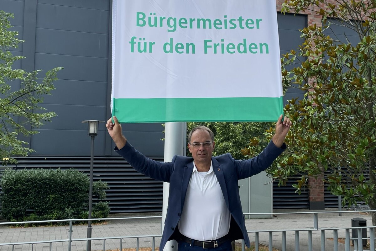 Ein Mann im Anzug h&auml;lt &uuml;ber sich eine gro&szlig;e Flagge mit dem Schriftzug "B&uuml;rgermeister f&uuml;r den Frieden".