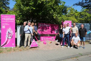 Bild vergr&ouml;&szlig;ern: Personen, Frauen und M&auml;nner stehen mit Spaten neben einem Sandhaufen, aus dem ein Glasfaserkabel heraus schaut. Neben und hinter ihnen Werbebanner der Telekom.