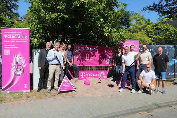 Personen, Frauen und M&auml;nner stehen mit Spaten neben einem Sandhaufen, aus dem ein Glasfaserkabel heraus schaut. Neben und hinter ihnen Werbebanner der Telekom.