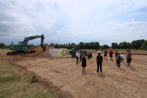 Bild vergr&ouml;&szlig;ern: Eine flache, ausgehobene Sandgrube, auf der zwei gro&szlig;e Bagger stehen. Im Kreis stehen dort mehrere Personen, die sich das Bauprojekt beschreiben lassen.