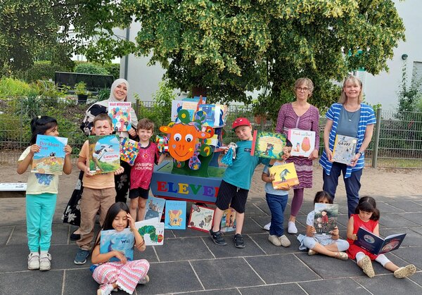 Kinder und drei Erzieherinnen stehen und sitzen in einer Gruppe, die halten B&uuml;cher in den H&auml;nden. Zwischen ihnen steht ein bunt bemaltes Schaf aus Holz.