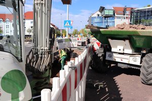 Bild vergr&ouml;&szlig;ern: Eine mit Warnbaken abgesperrte Baustelle. Dort steht ein Bagger sowie ein Fahrzeug mit aufgeladener Erde, ein Mann mit Schaufel arbeitet.