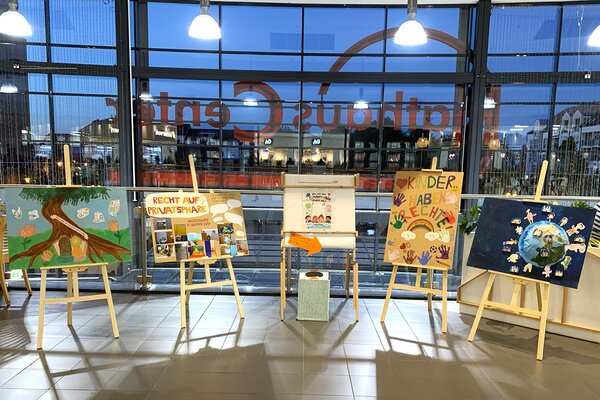 Auf vier Staffeleien stehen gro&szlig;e Bilder und Plakate. Sie sind bunt gestaltet und zeigen beispielsweise Sprechblasen, Handabdr&uuml;cke oder Naturmalereien.