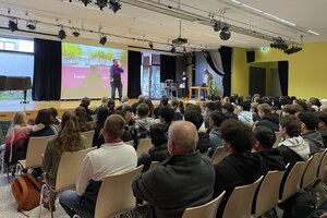 Bild vergr&ouml;&szlig;ern: Ein Saal mit B&uuml;hne und Pr&auml;sentationsleinwand. Auf der B&uuml;hne steht ein Mann mit Mikrofon, im Raum sitzen Jugendliche auf St&uuml;hlen und h&ouml;ren ihm zu.